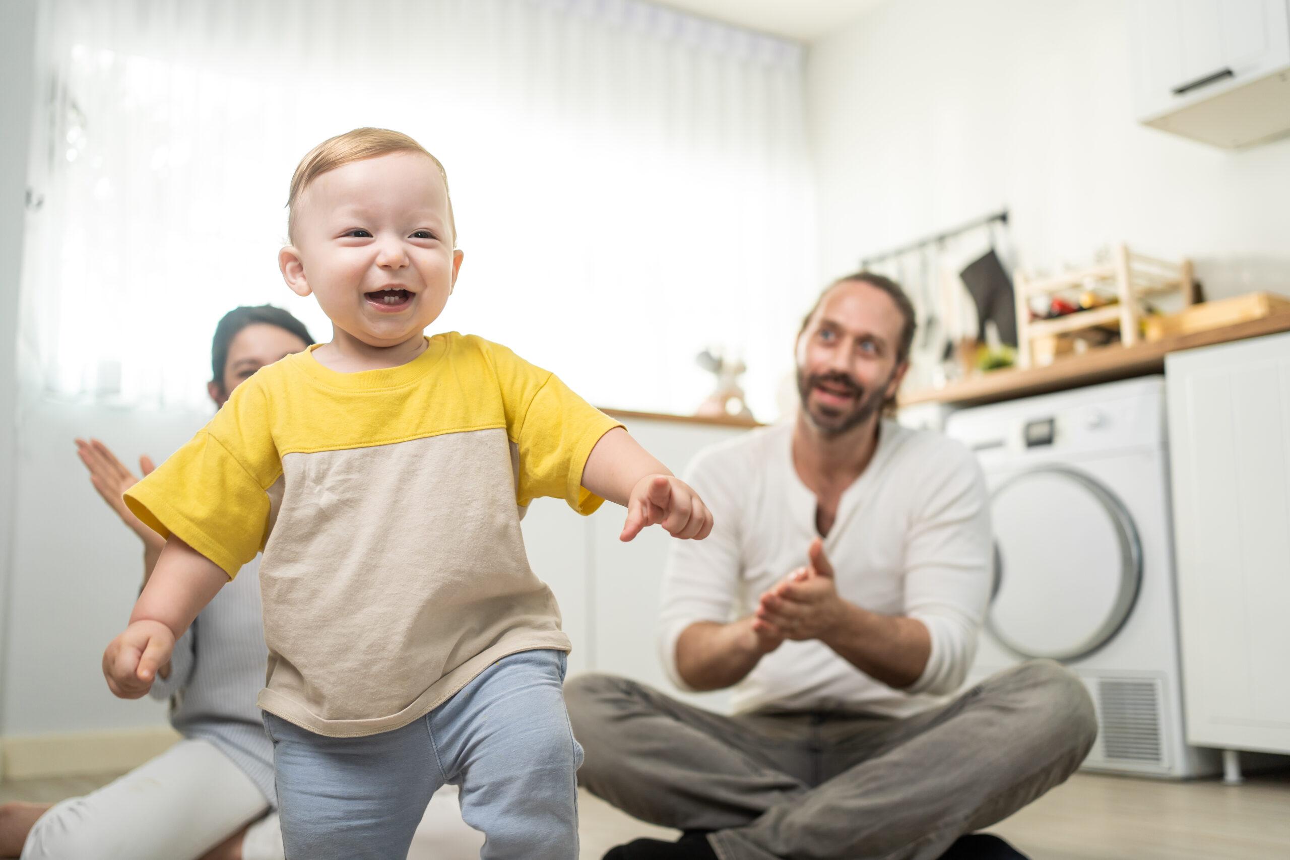 Caucasian baby boy child learn to walk with parents support in house. Happy family, mother and father helping young toddler son taking first step walk on floor to develop skill in living room at home.;